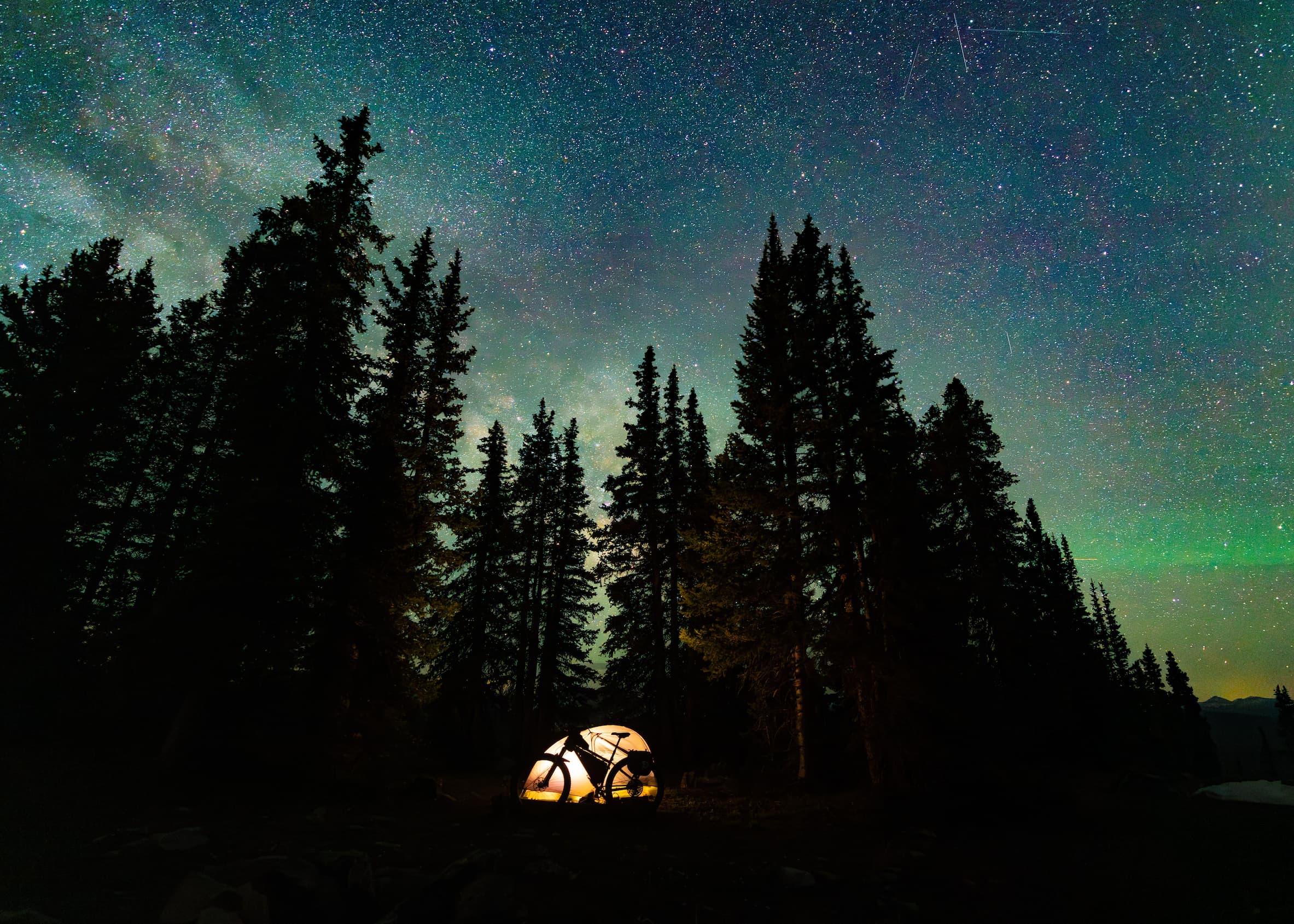 A glowing tent is set up among tall pine trees under a star-filled night sky with visible constellations and a hint of the Milky Way.