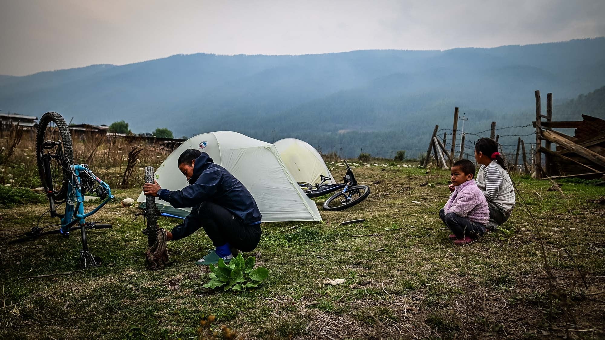 Person sitting on the ground next to a tent with two children sitting nearby, mountains in the background.