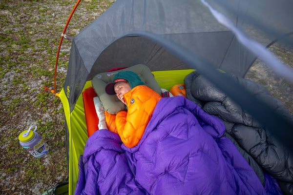 A person in a bright orange jacket and green hat is sleeping inside a tent, wrapped in a purple sleeping bag. Camping gear and a water bottle are visible nearby on grassy ground.