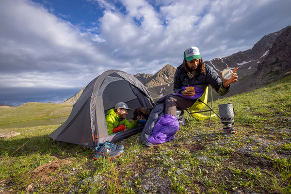 Two people sit by a tent in a grassy mountain landscape; one prepares food on a portable stove while the other is inside the tent with a dog. The sky is partly cloudy and the scene is peaceful and outdoorsy.