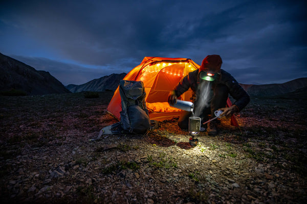 A person wearing a headlamp sits by an orange tent with string lights, preparing food on a camp stove at dusk in a rocky mountain landscape. A backpack is placed beside the tent.