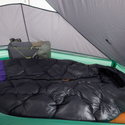 A cozy black sleeping bag lies inside a green Big Agnes String Ridge VST 1.5 tent, with a gray backpack and gear by the back wall. Soft light filters through the mesh ceiling and walls of this ultralight shelter.